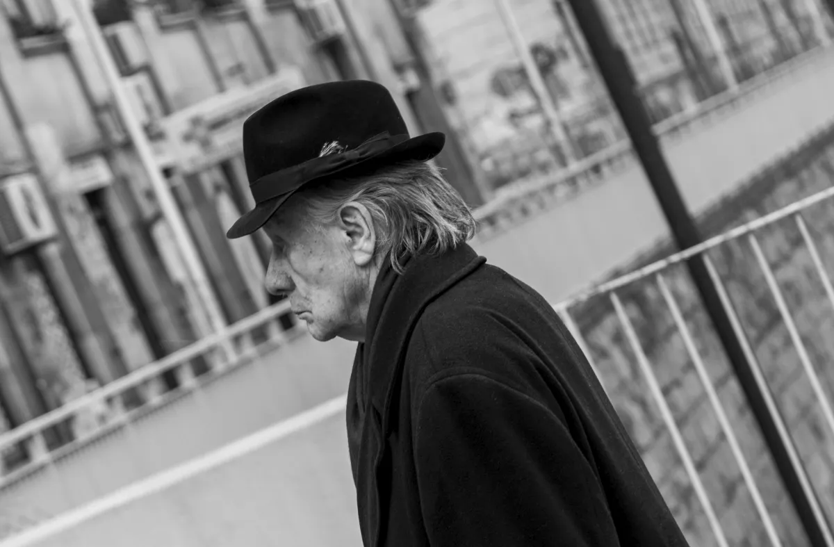 Older man in a dark fedora and heavy coat walking in side profile beside bridge railings